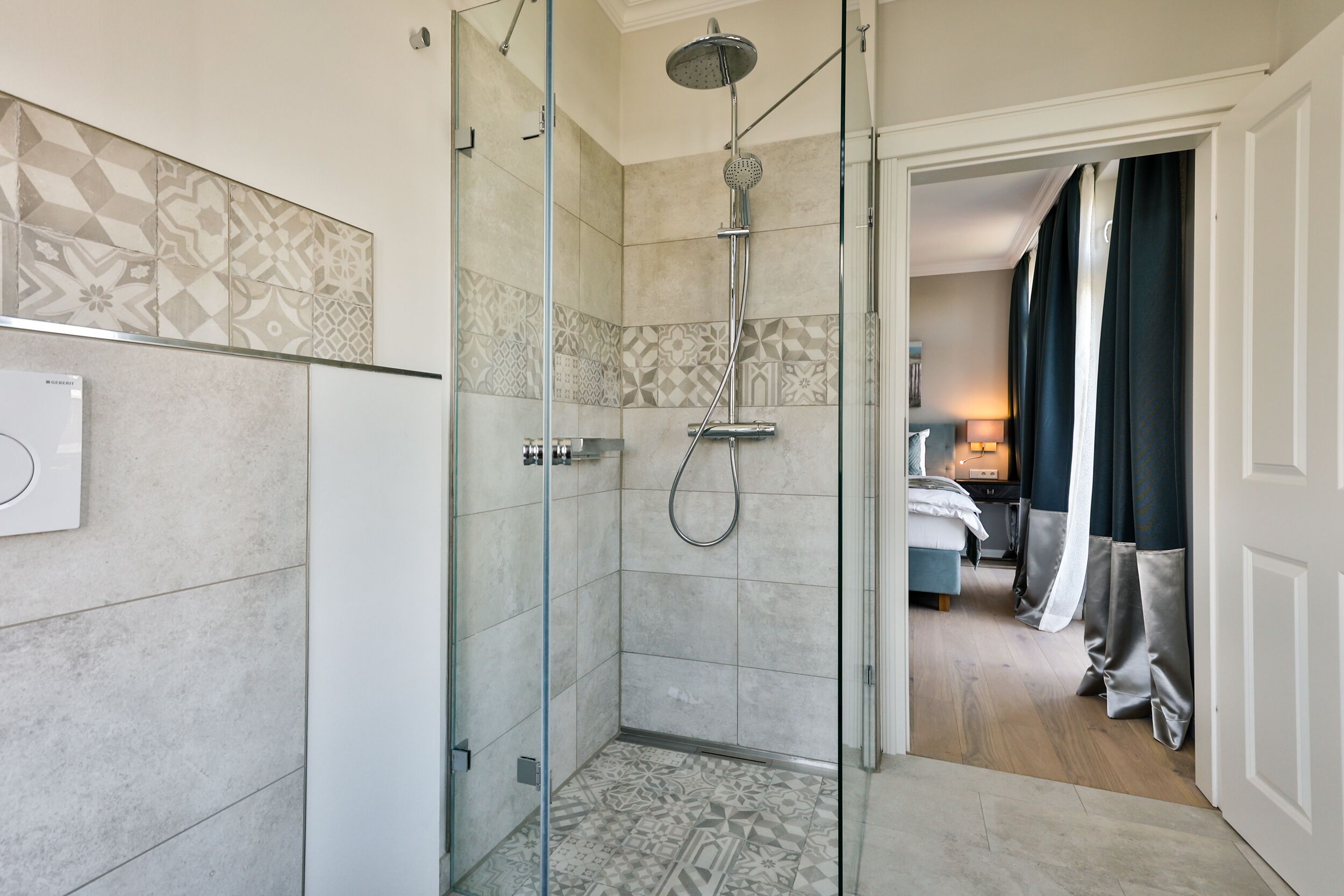A modern bathroom with a glass-enclosed shower, patterned tiles, and a visible bedroom with a bed and curtains.