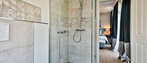 A modern bathroom with a glass-enclosed shower, patterned tiles, and a visible bedroom with a bed and curtains.