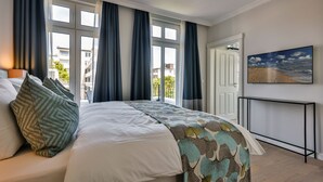 Une chambre avec un grand lit, des rideaux sombres, une vue sur les bâtiments à l’extérieur et un tableau accroché au mur.