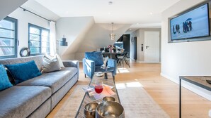 A modern living room with a grey sofa, a coffee table with decorative items, a dining area with a blue chair, and a TV mounted on the wall.