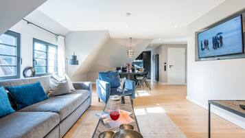 A modern living room with a grey sofa, a coffee table with decorative items, a dining area with a blue chair, and a TV mounted on the wall.