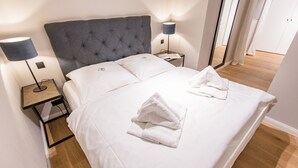 A neatly made bed with white linens and two towels, a grey upholstered headboard, and bedside lamps on matching nightstands.