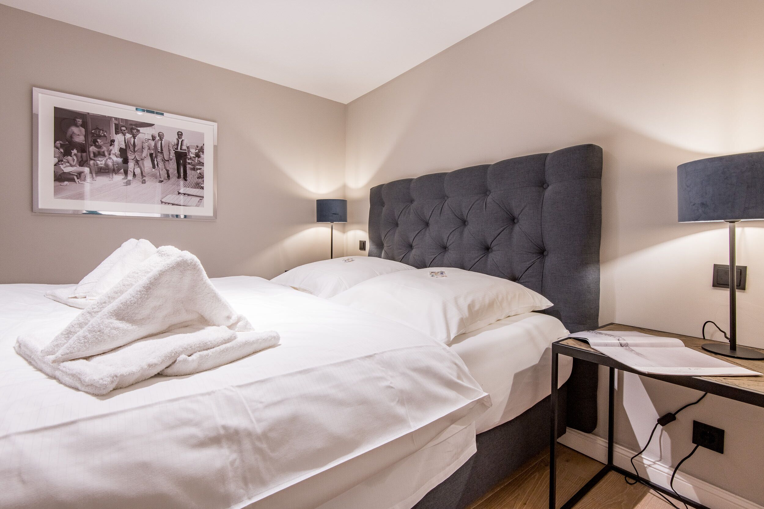 Hotelkamer met een groot bed, twee bedlampjes, een ingelijst foto aan de muur en een kleine tafel met lamp.