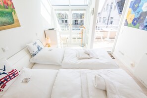 A bright room with two beds, a painting on the wall, and a view of buildings outside.