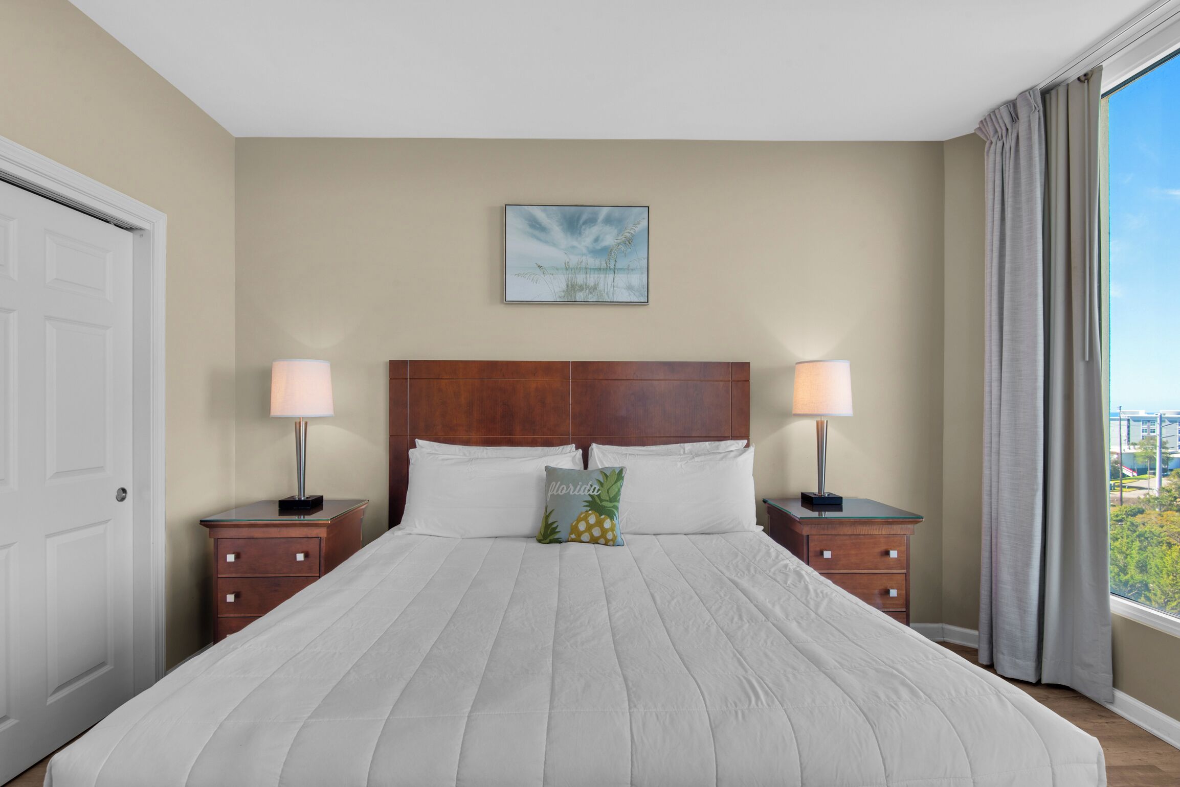 A hotel room with a large bed, two bedside tables with lamps, a wooden headboard, a framed picture on the wall, and a window with curtains.