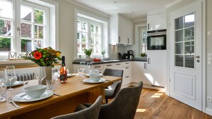 Een moderne keuken met witte kastjes, een houten eettafel voor vier personen en een groot raam dat natuurlijk licht binnenlaat.