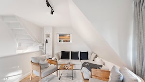 Una moderna camera da letto mansardata con soffitto inclinato, una scala a scomparsa, un’accogliente area salotto con divano e poltrone, e un’ampia finestra con vista sulla città.