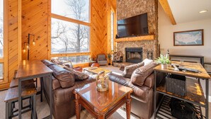 A cozy cabin interior with a stone fireplace, leather sofas, a wooden dining table, and large windows offering a view of the outdoors. - Epic Wisp Views w/ Fireplace, Game Room & Hot Tub (McHenry)