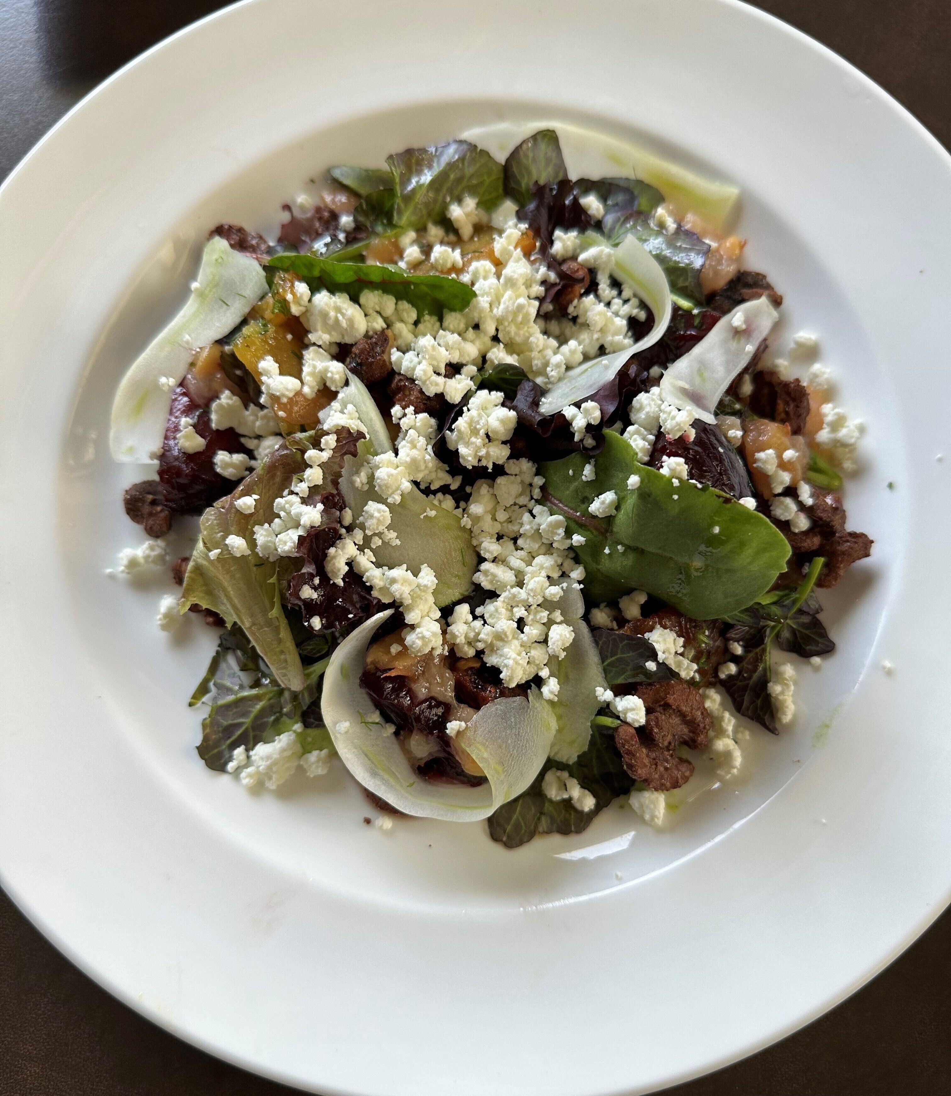Un plato blanco con una ensalada de vegetales, queso y posiblemente carne.