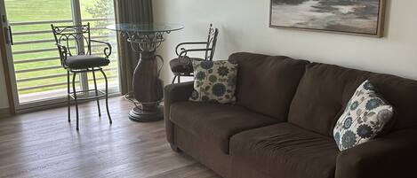 A living room with a brown sofa, two chairs, a glass-top table, and a painting on the wall.