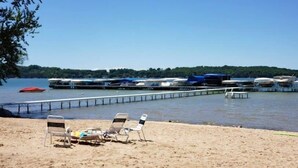 En strand med solstolar, en brygga som sträcker sig ut i vattnet och båtar som ligger förtöjda.