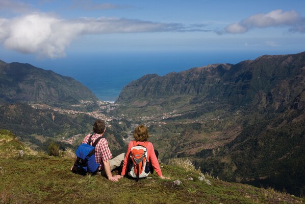 Dos personas sentadas en una colina cubierta de hierba, con vistas a un valle, al océano y a las montañas.