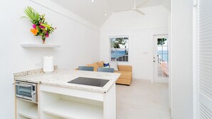 Ein heller, weißer Innenraum mit Küchenzeile, einschließlich Arbeitsfläche, Ofen und Deckenventilator. Im Hintergrund ist ein beiges Sofa zu sehen, und ein Fenster bietet Blick ins Freie.