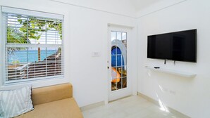 Ein Zimmer mit einem beigefarbenen Sofa, einem an der Wand montierten Fernseher, einem Fenster mit Jalousien und einer Tür mit Blick ins Freie.