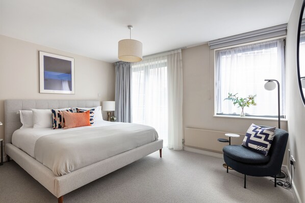 A neatly arranged hotel room with a large bed, a side table, a chair, and a window with curtains.