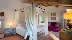 Quarto com uma cama de dossel, mesa de cabeceira com abajur, cômoda de madeira e sofá roxo.