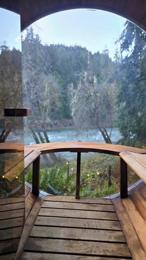Een houten terras met glazen hekwerk met uitzicht op een rivier en een bebost gebied.
