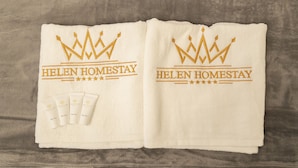Una toalla con el texto "HELEN HOMESTAY" y un logotipo de corona, junto con cuatro pequeños tubos etiquetados como "Loción Corporal", "Jabón Líquido Corporal", "Champú" y "Acondicionador".