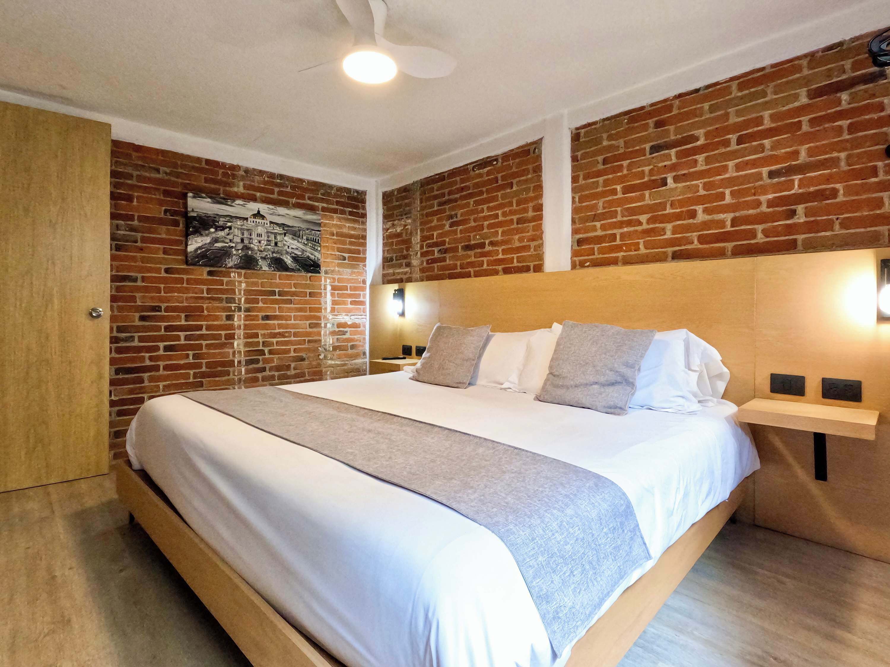 Een hotelkamer met een bakstenen muur, een bed met wit beddengoed, een houten hoofdeinde en een nachtkastje met een lamp.