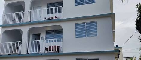 A multi-story residential building with balconies, a car parked in front, and a palm tree in the background.