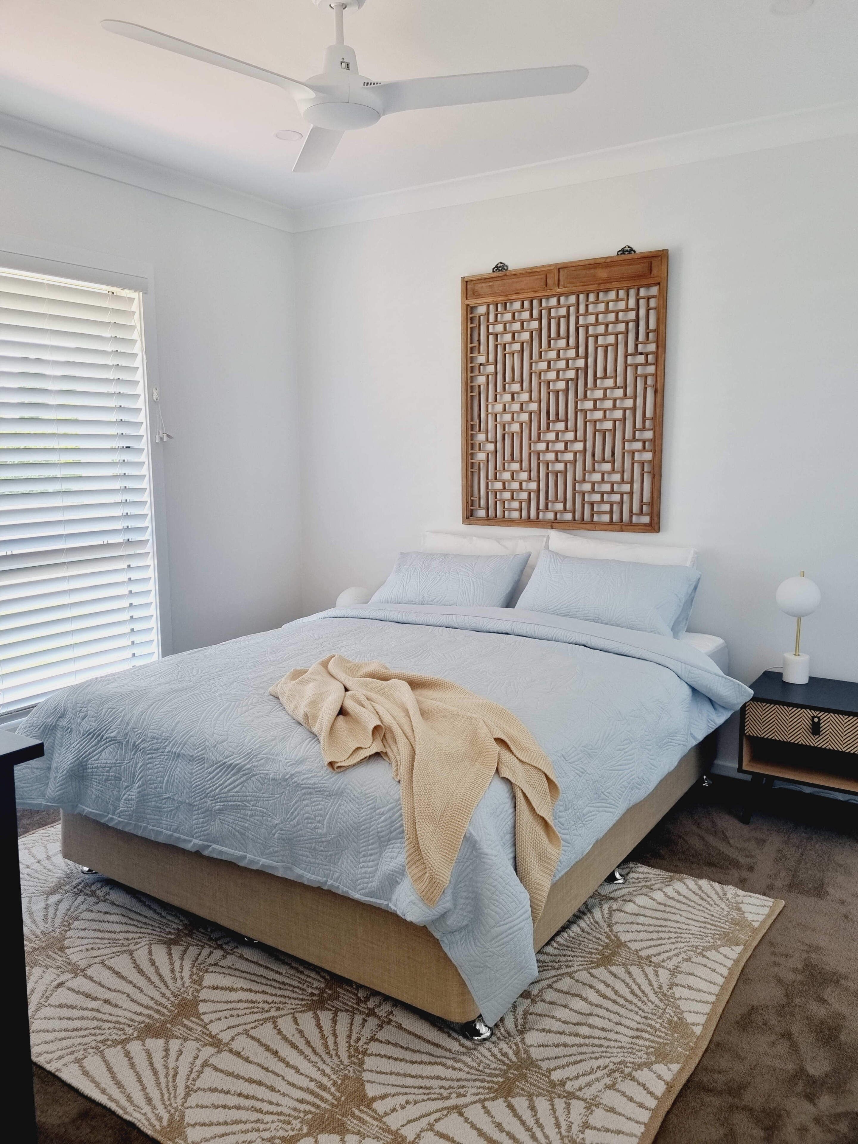 Une chambre à coucher comprenant un lit, un ventilateur de plafond, un panneau décoratif fixé au mur et une table de chevet avec une lampe.