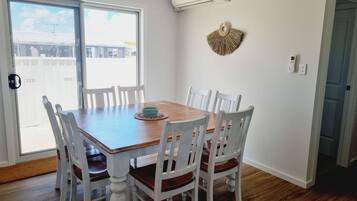 A dining area with a wooden table and white chairs, an air conditioning unit, and a decorative wall piece.
