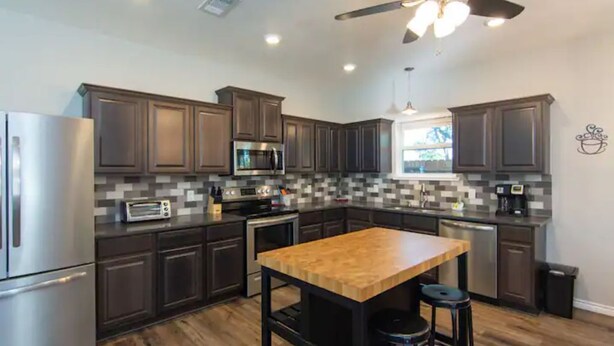 A modern kitchen with dark wood cabinets, stainless steel appliances, a wooden island with stools, and a ceiling fan.