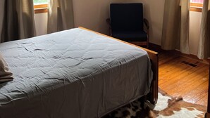 Ein Schlafzimmer mit einem Bett, einem Sessel und einem Kuhfellteppich. Es gibt zwei Fenster mit Vorhängen, einen Deckenventilator und Wandbilder.