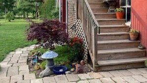 Ein Haus mit einer Holztreppe, die zu einer roten Tür führt, einem Garten mit verschiedenen Pflanzen und einem farbenfrohen, gestreiften Sonnenschirm.