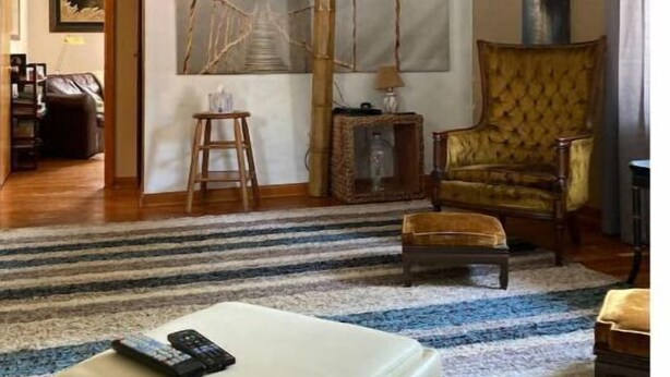 Ein Wohnzimmer mit einem großen Flachbildfernseher an der Wand, einem gelben Sessel, einem Holztisch und einem Streifenmusterteppich.