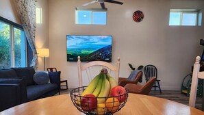 A living room with a wooden table, a TV on the wall, a ceiling fan, and a window with curtains. - The Gregory: 2 Bed/1 Bath Guesthouse (Phoenix)