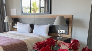 A hotel room with a bed, a bedside table with a lamp, a vase with red roses, and a window with a view of palm trees. - theGREEN Villa, your solar powered home away from home (Cape Town)