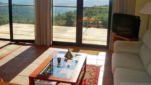 Ein Wohnzimmer mit einer Couch, einem Couchtisch und einem Fernseher. Das Zimmer hat große Fenster mit Blick auf Berge.
