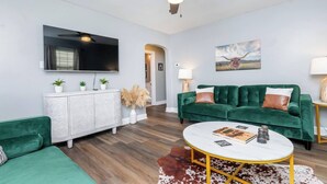 Un salon moderne avec un canapé vert, un meuble blanc, un téléviseur à écran plat, un ventilateur de plafond et une table basse ronde.