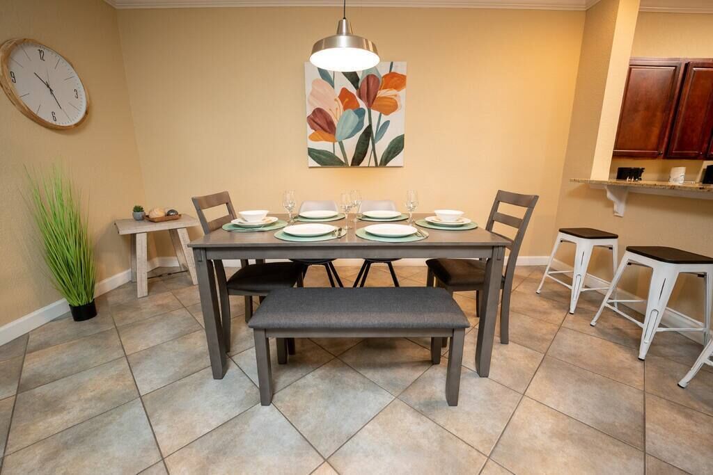 Un coin repas avec une table dressée pour quatre personnes, un banc, une horloge et un tableau accroché au mur.
