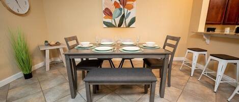 Un coin repas avec une table dressée pour quatre personnes, un banc, une horloge et un tableau accroché au mur.
