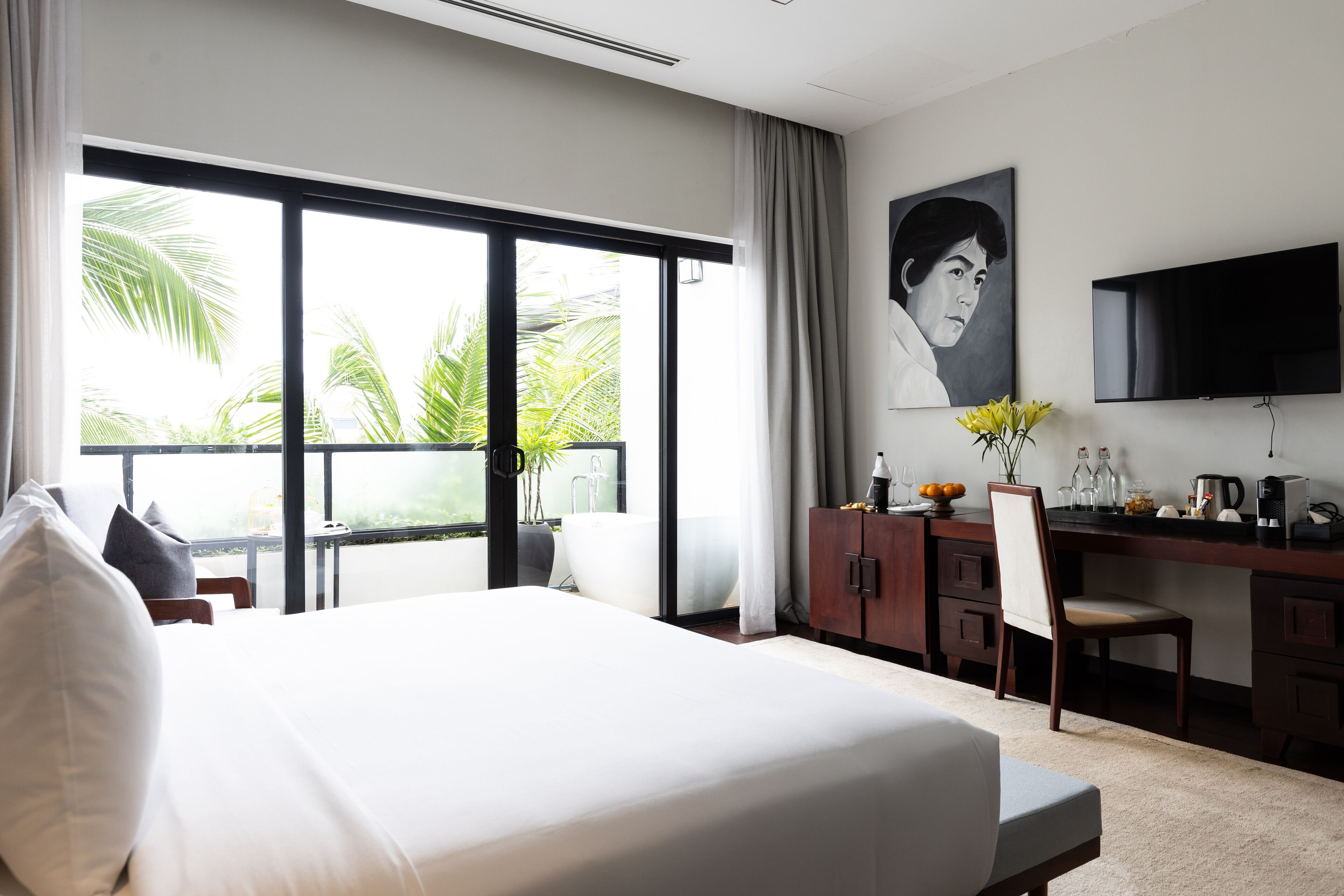 A hotel room with a large bed, a desk with a chair, a television, and a window with a view of palm trees.