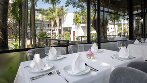 Ein Restaurant mit weißen Tischtüchern, grau gepolsterten Stühlen und Blick auf Palmen und Gebäude durch große Fenster.
