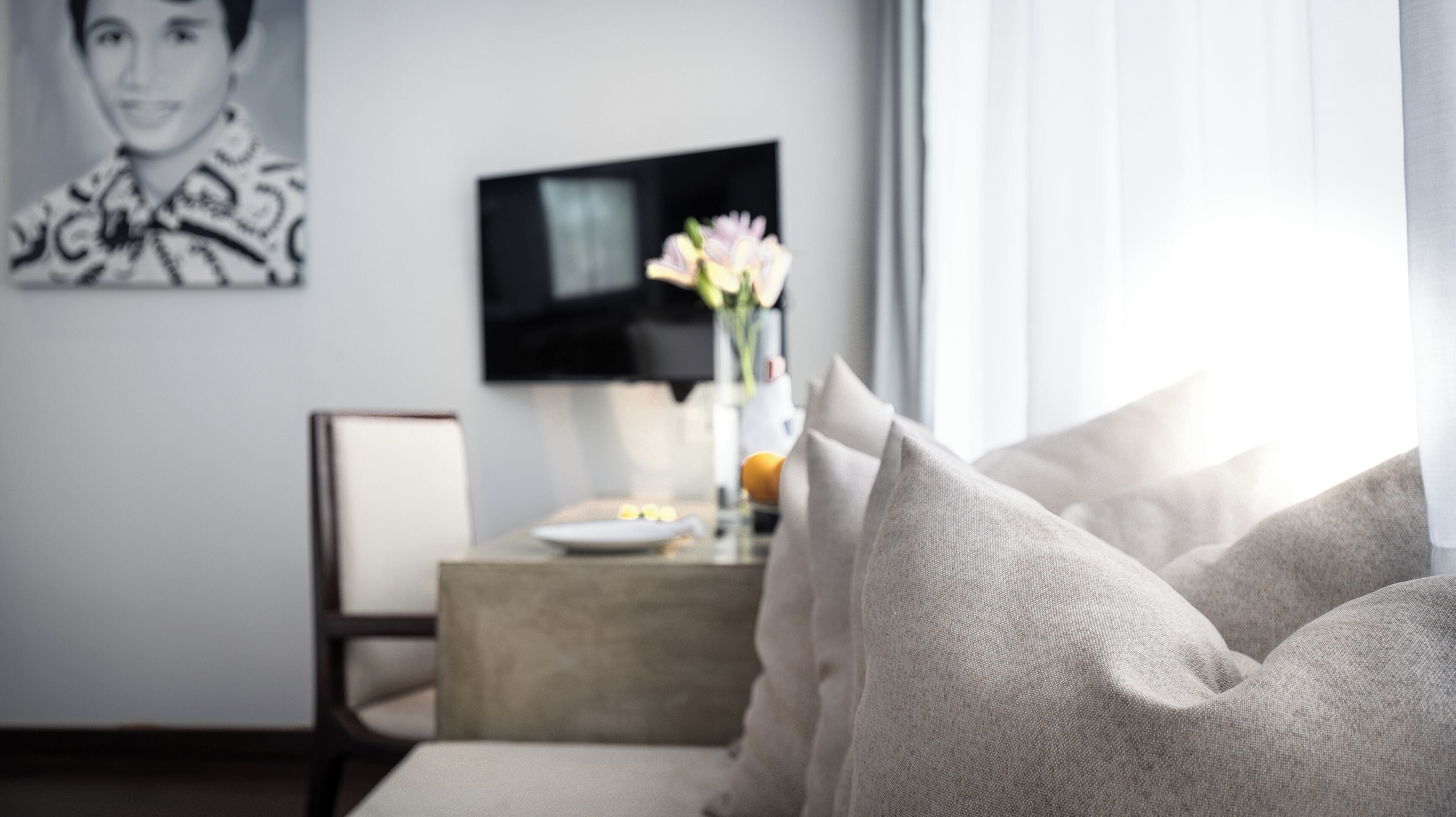 A modern living room with a sofa, a flat-screen TV, a vase with flowers, and a dining table with a plate of fruit.