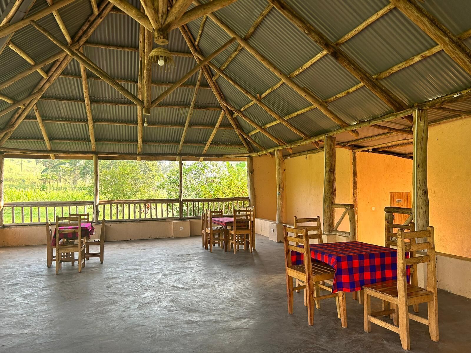 A covered outdoor seating area with wooden tables and chairs, a corrugated metal roof, and a view of greenery outside.