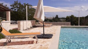 A swimming pool with lounge chairs and umbrellas, surrounded by stone walls and greenery. - Trullo Genista with Private Pool in the Golden Triangle, Puglia Italy (Martina Franca)