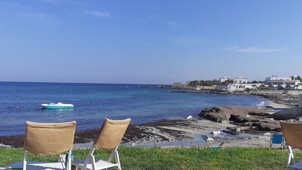 A seaside view with chairs facing the ocean, rocky shoreline, and buildings in the background. - Trullo Genista with Private Pool in the Golden Triangle, Puglia Italy (Martina Franca)