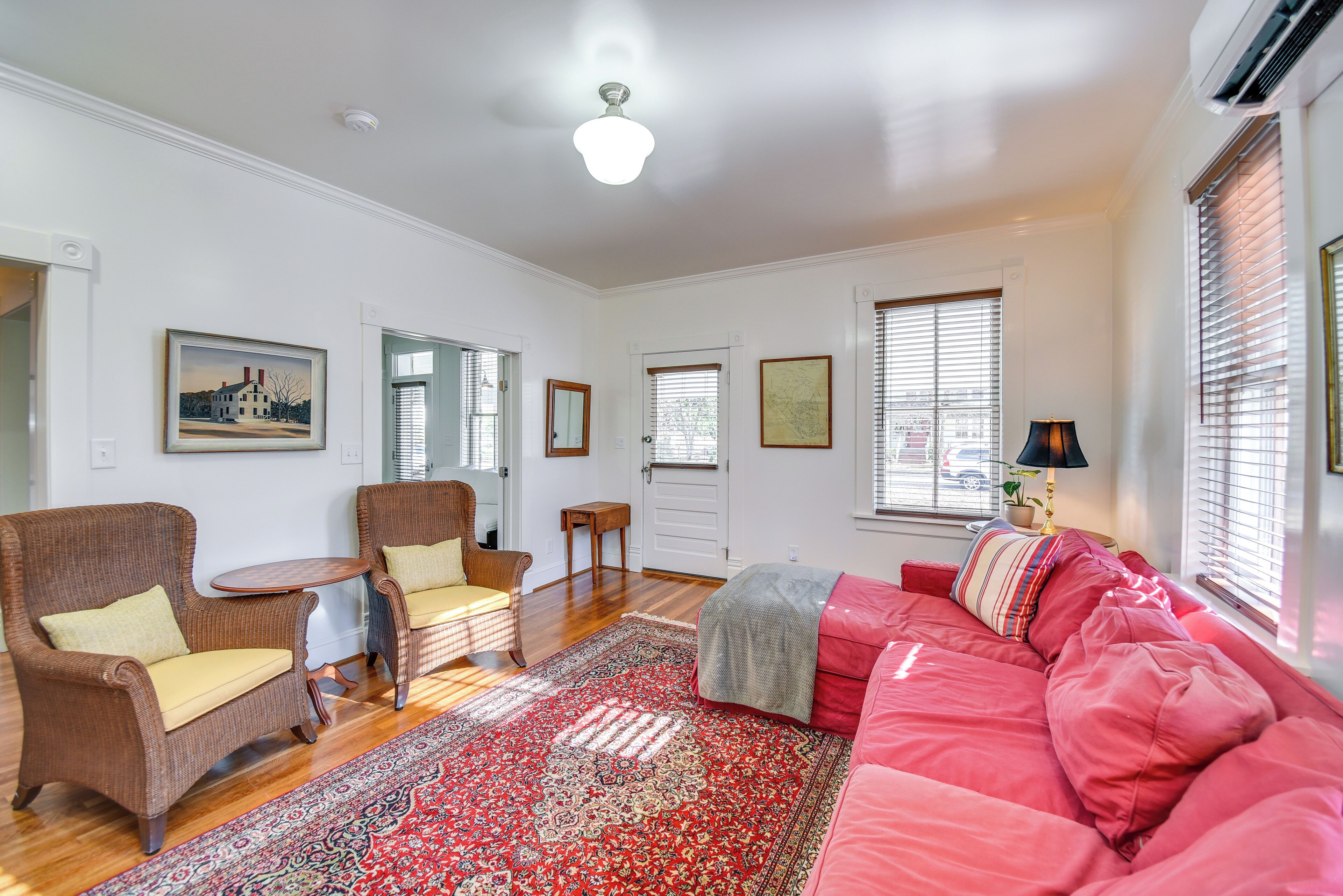 A spacious living room with a large red sofa, two wicker chairs, a small round table, and a patterned rug.