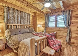 Une pièce de cabane en bois comprenant un lit, une petite table avec une bouteille et des verres, un ventilateur de plafond et des rideaux à motifs.