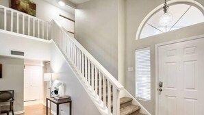 Un escalier avec des rampes blanches menant à un niveau supérieur, une porte avec une fenêtre, et une petite table avec des livres et une lampe.