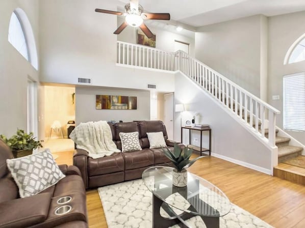 A spacious living room with a brown leather sofa, a glass coffee table, and a white staircase.