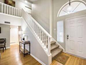 A staircase with white railings leading to an upper level, a door with a window, and a small table with books and a lamp.