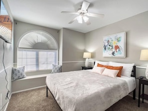 A bedroom with a bed, a ceiling fan, a window with blinds, a nightstand with a lamp, and a wall-mounted artwork. - Chandler 3BR | Pool + Game Room | Pet Friendly (Chandler)