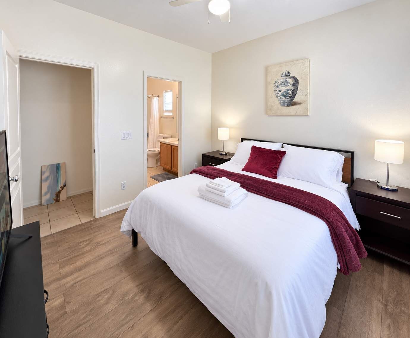 A hotel room with a bed, bedside tables, a painting, and a bathroom visible through an open door.