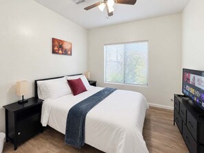 Ein Schlafzimmer mit einem Bett, einem Nachttisch, einem Fernseher, einem Deckenventilator, einem Fenster mit Jalousien und einem an der Wand hängenden Kunstwerk.